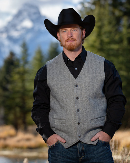 Man wearing a cowboy hat, gray vest, and black shirt in a natural setting with mountains and trees.