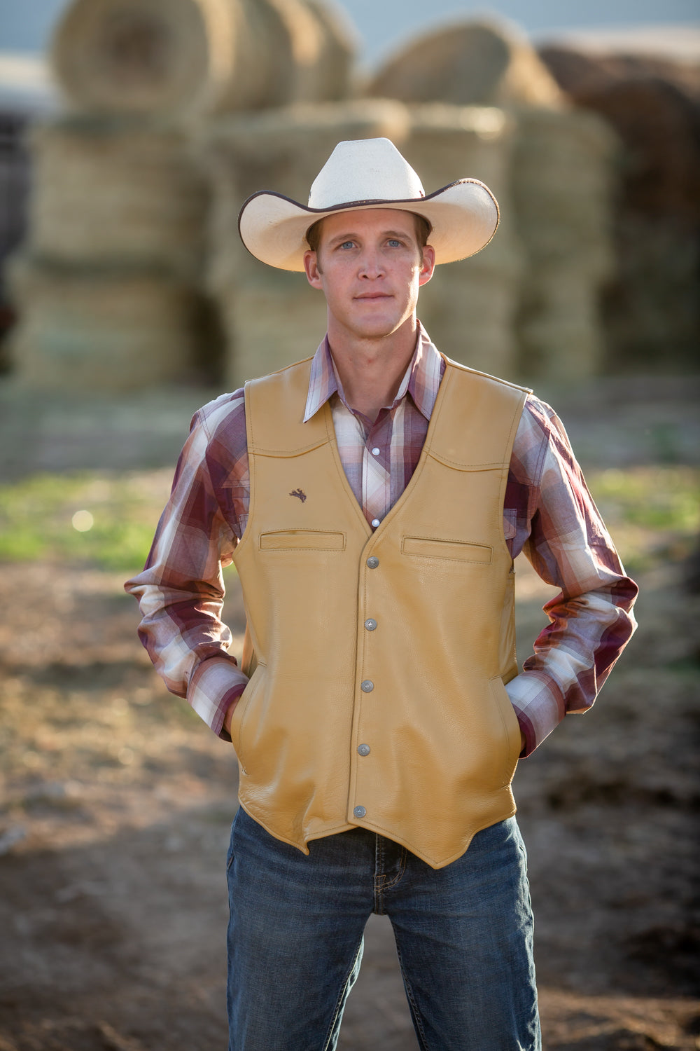 Man wearing a cowboy hat, yellow vest, and plaid shirt standing in front of hay bales.