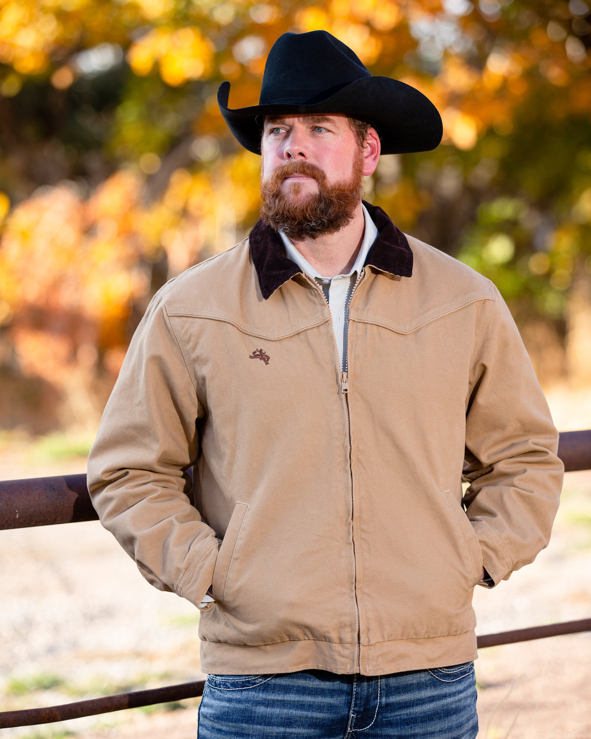 Man wearing a tan jacket and black cowboy hat standing outdoors with autumn trees in the background